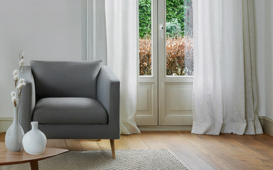 Modern eingerichtetes Wohnzimmer mit grauem Sessel, weißen Vorhängen aus JOKA Dekostoffen und heller Holzoptik.