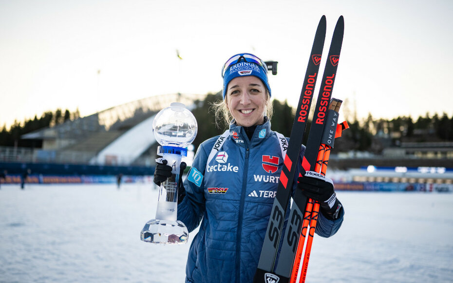 JOKA Biathletin Franziska Preuß strahlt nach dem Wettkampf mit Skiern und einer Trophäe in der Hand vor winterlicher Kulisse.