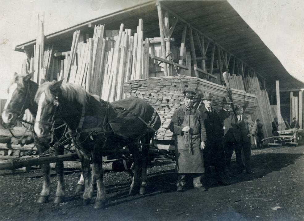 Schwarz-weoß Foto einer alten Scheune mti vielen Hölzern, davor ist eine Kutsche mit Pferden zu sehen, die das Holz transportieren.