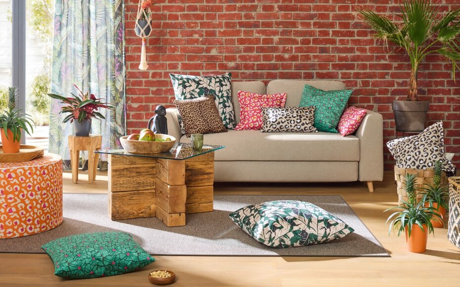 Wohnzimmer mit JOKA Möbelstoffen: Beige Couch vor roter Backsteinwand, dekoriert mit farbenfrohen Kissen in Tier- und Pflanzenmustern. Dazu Holztisch mit Glasplatte, grüne Pflanzen und Vorhänge mit Blättermotiv.