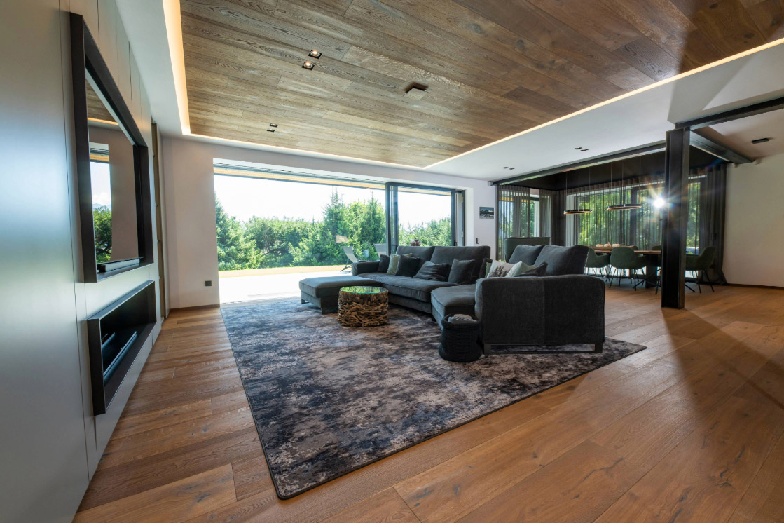 Ein stilvolles Wohnzimmer mit großem Fenster, das auf eine Terrasse führt. Der Parkettboden und die Holzdecke schaffen ein harmonisches und gemütliches Ambiente.