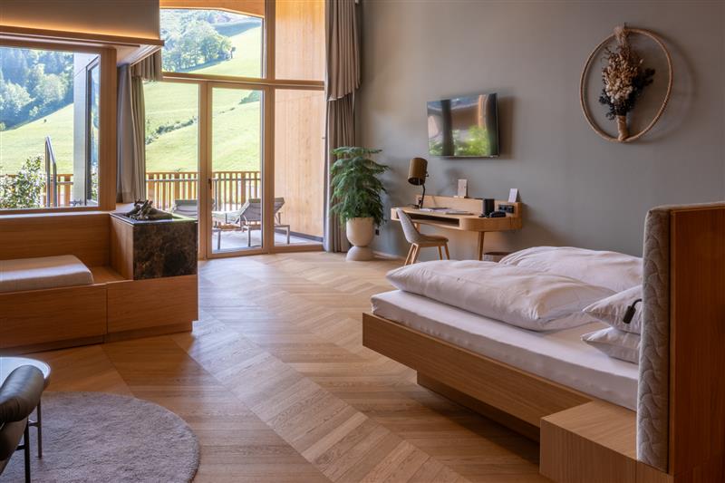 Modern eingerichtetes Hotelzimmer mit großzügigem Panoramafenster und Balkonblick auf eine grüne Hügellandschaft. Der Boden ist mit einem eleganten Fischgrätparkett aus hellem Holz ausgestattet, das harmonisch mit der natürlichen Einrichtung aus Holz und sanften Farben abgestimmt ist.
