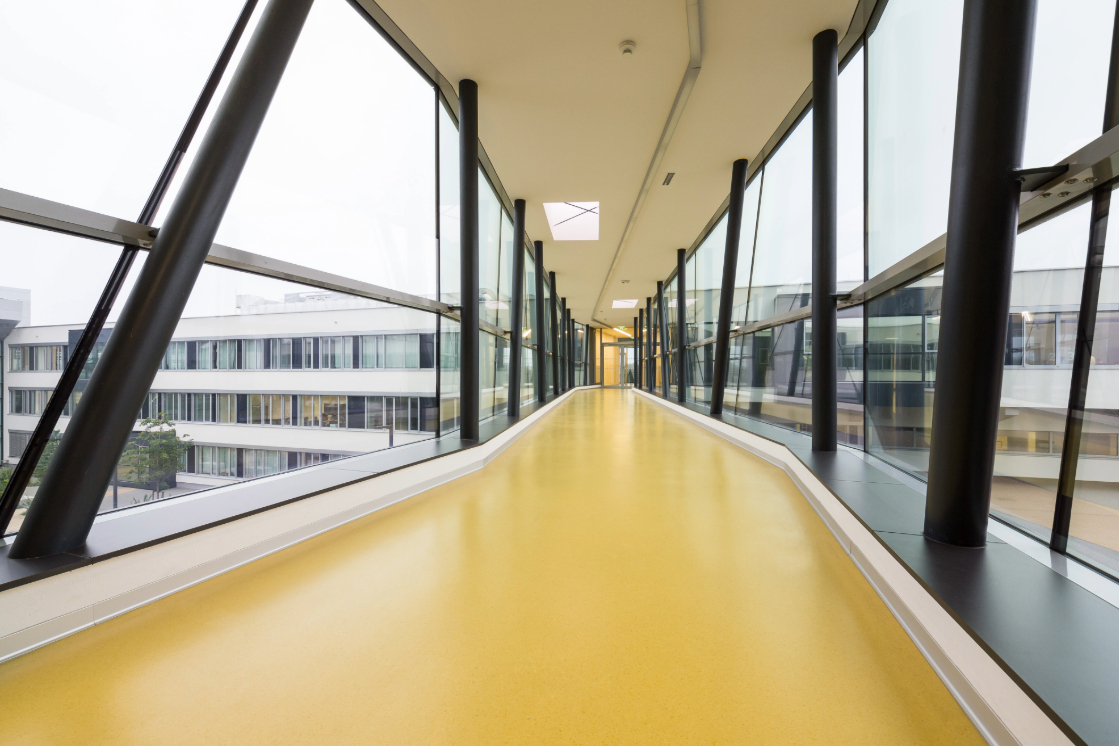 Ein langer, verglaster Übergang mit einem gelben Bodenbelag verbindet zwei moderne Gebäude. Große Fenster an beiden Seiten lassen viel Licht herein und bieten einen klaren Blick auf die Außenfassaden.